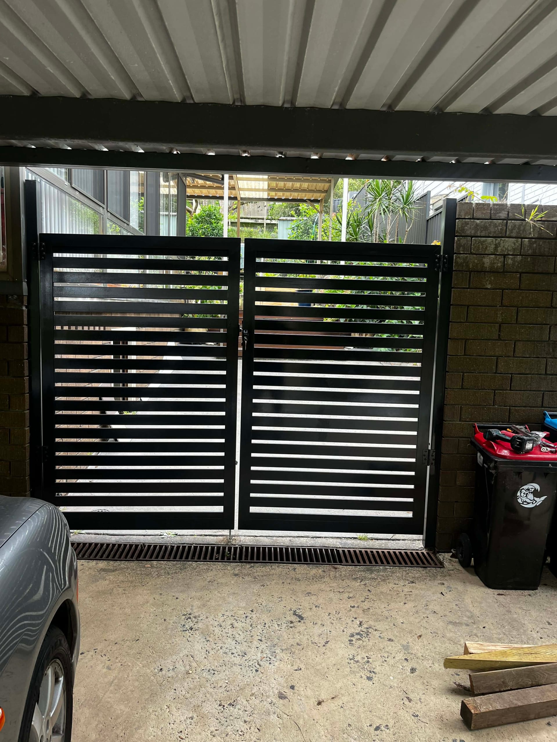 Pair of black aluminium horizontal slat gates closed across a carport opening between a brick wall and a besser-block wall