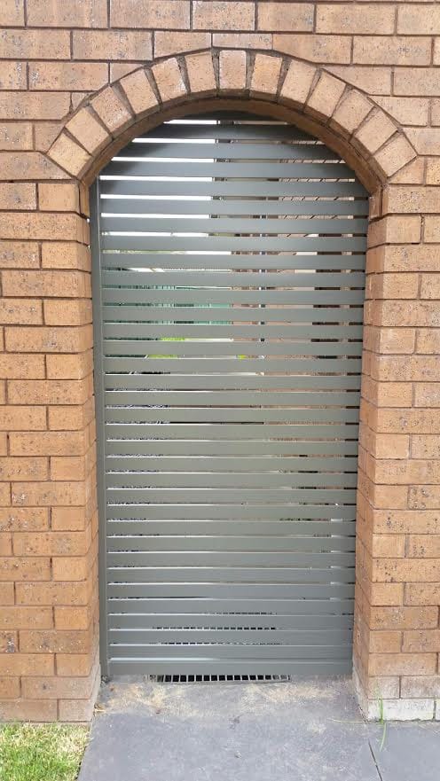 Grey-green aluminium horizontal slat gate fitted into an arched brick opening in a face-brick wall