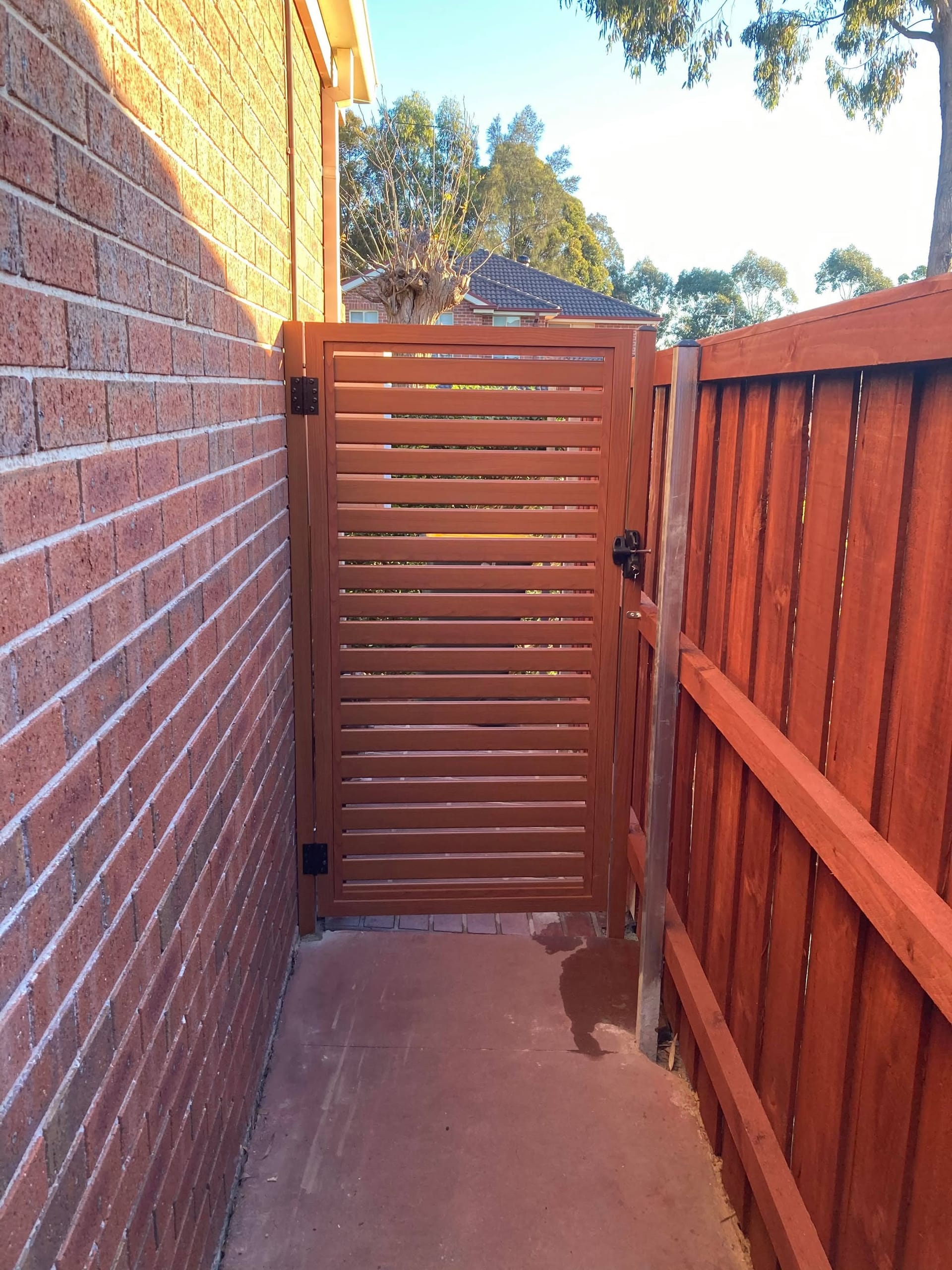 Timber-look woodgrain aluminium horizontal slat pedestrian gate installed between a brick wall and a stained timber paling fence
