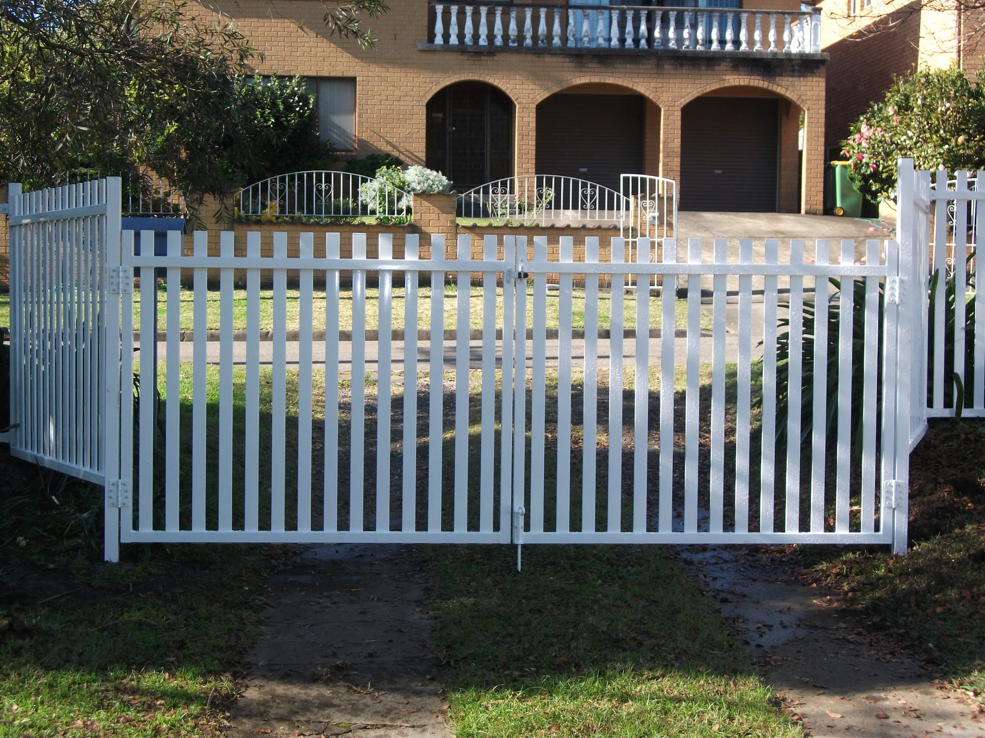 White aluminium vertical picket double gates across a driveway with matching picket fencing either side of a brick home