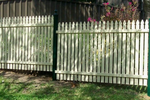 Cream aluminium vertical picket fencing with a green post and spear-top pickets in front of a dark paling fence