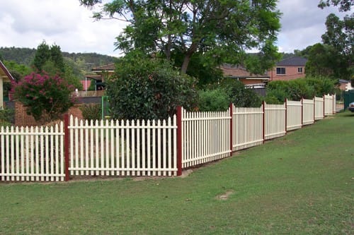 Cream aluminium vertical picket fencing with red posts stepping down a grassed slope in front of a brick house