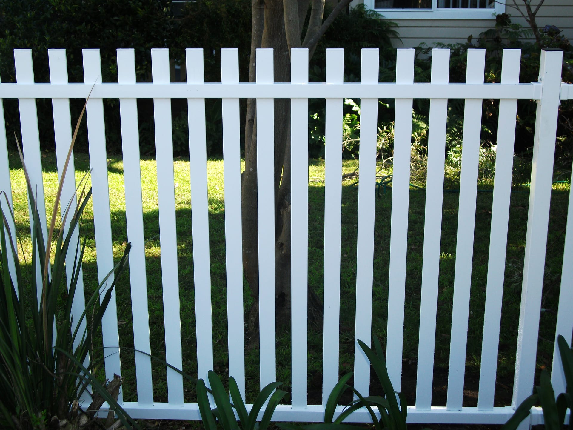White aluminium vertical picket fencing with flat-top pickets running along a garden boundary in front of a lawn and a tree